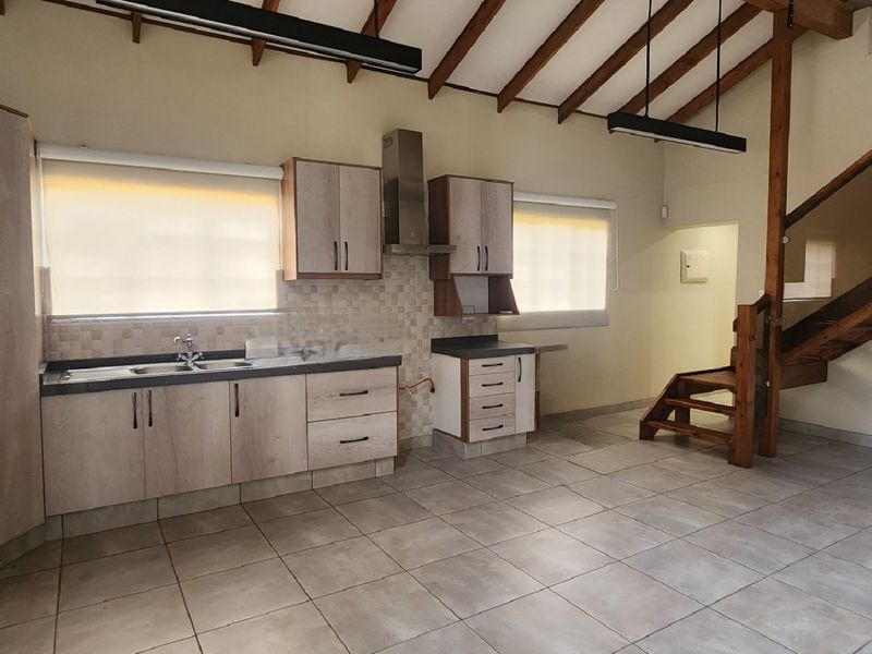 Open-plan kitchen area featuring light wood cabinetry, tiled flooring, and a striking timber staircase with glass balustrade under a vaulted ceiling.