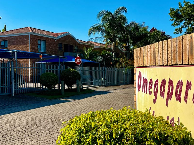 The entrance to Omegapark apartment complex featuring a paved driveway, brick boundary wall, and secure metal fencing with covered parking areas.