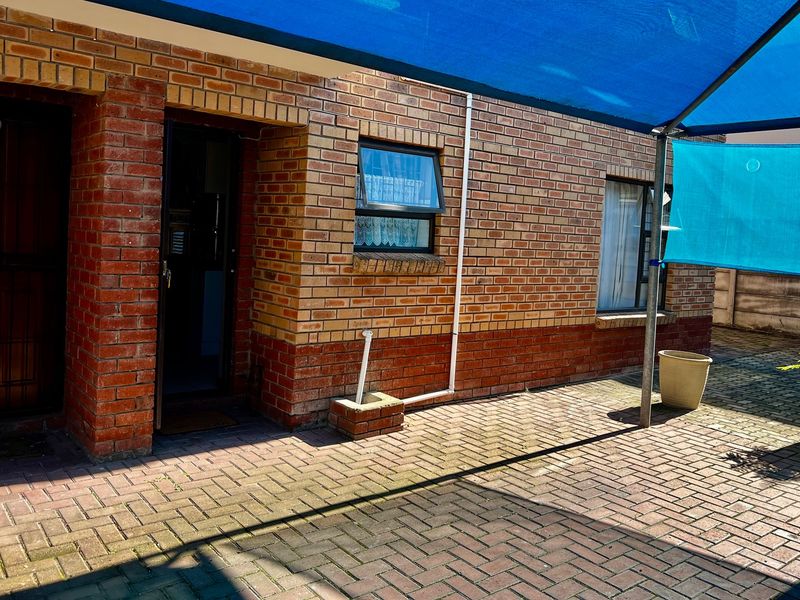 A paved outdoor patio area featuring a face brick facade, a blue shade sail, and a secure entrance door.