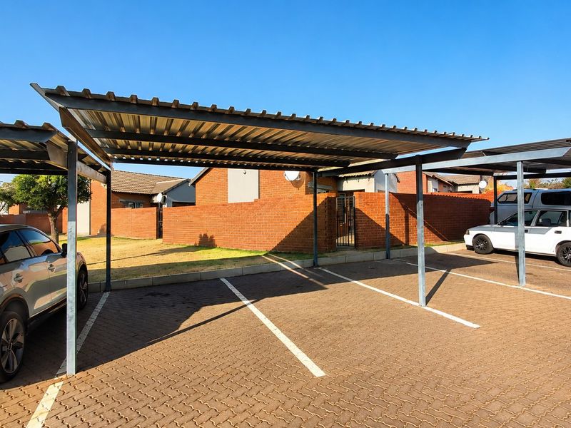 Covered carport parking area with paved driveway and brick boundary wall in a townhouse complex.