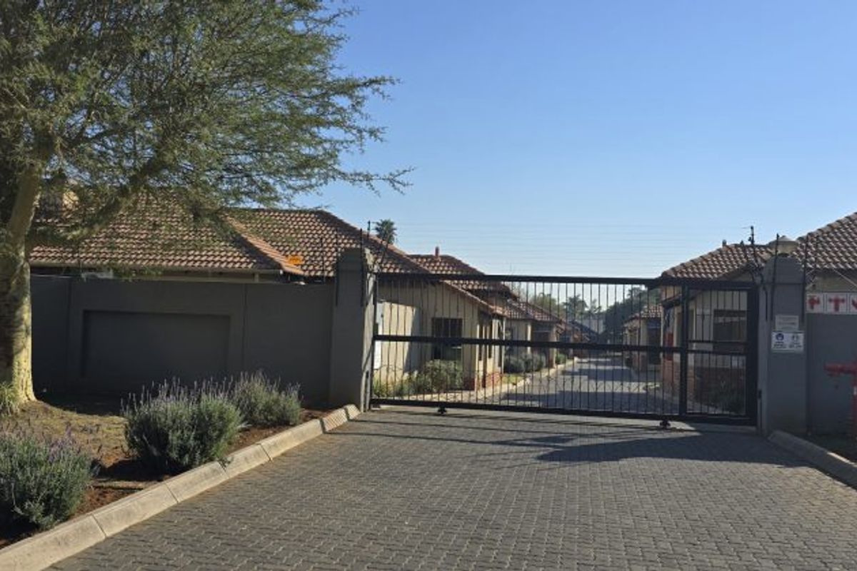 View of the secure gated entrance to the townhouse complex, featuring a paved driveway, perimeter walling with electric fencing, and tiled roof structures.