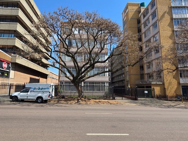 Street view of a multi-story apartment building complex with mature trees and security fencing along the sidewalk.
