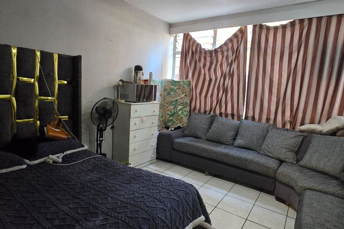 A bedroom featuring tiled flooring and a large window, currently used as a multi-purpose living and sleeping space.