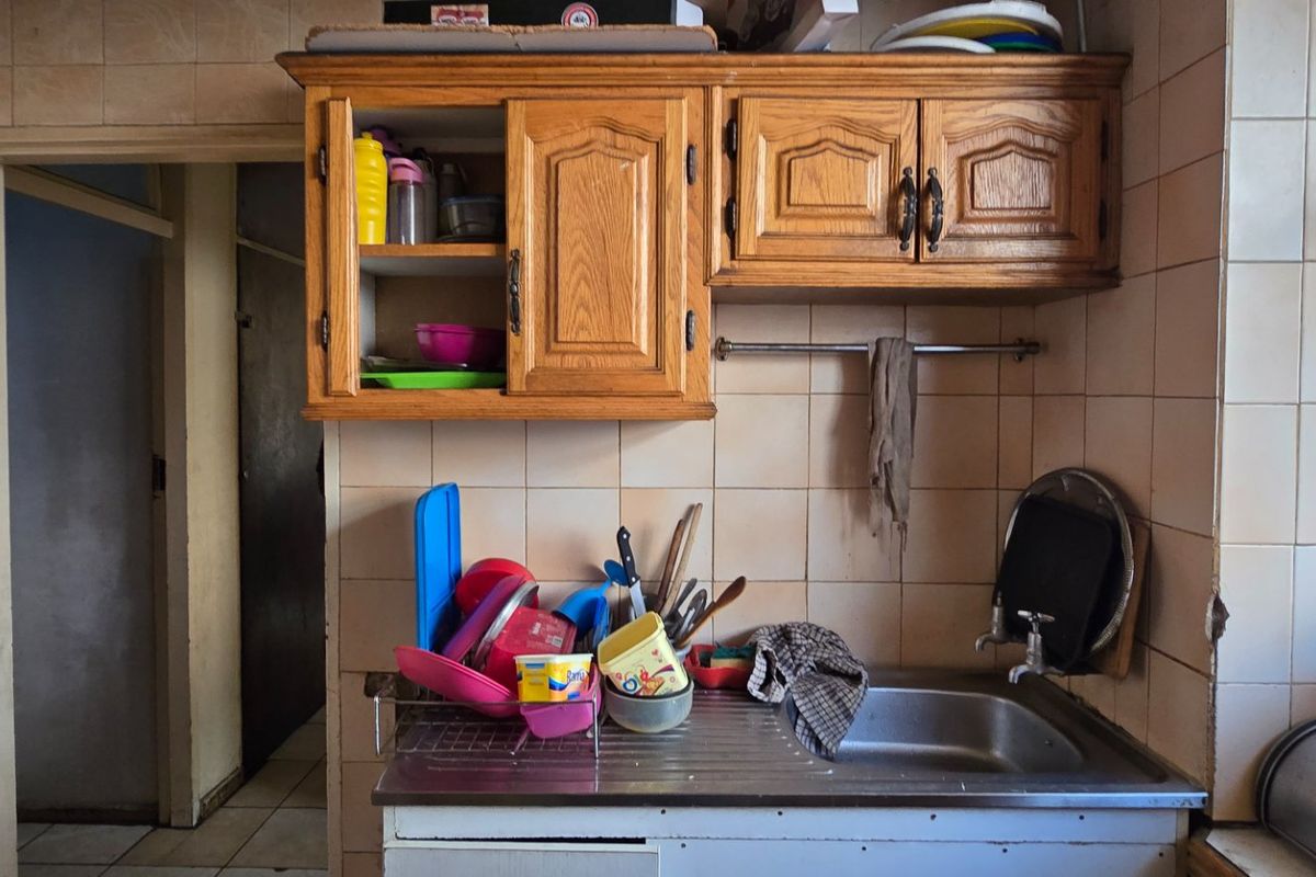 A dated kitchen area featuring wooden cabinetry, a stainless steel sink, and ceramic wall tiling, requiring cosmetic updates to improve its overall condition.