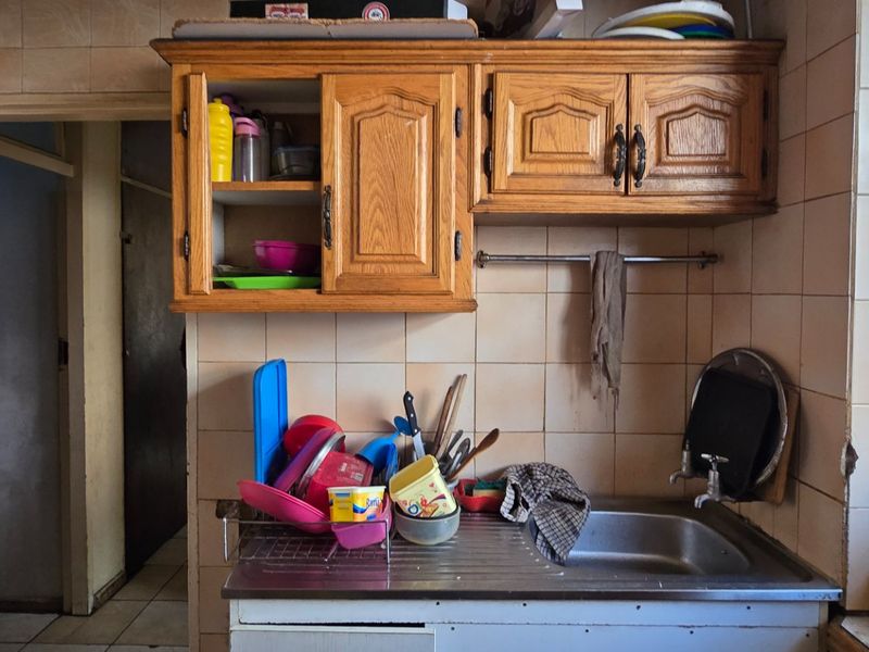 A dated kitchen area featuring wooden cabinetry, a stainless steel sink, and ceramic wall tiling, requiring cosmetic updates to improve its overall condition.