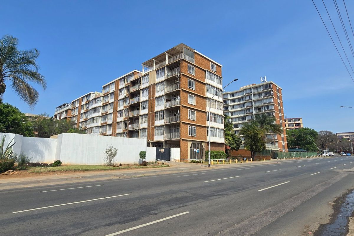 A street-level view of a multi-story apartment complex featuring brick facades and private balconies, situated along a main road.