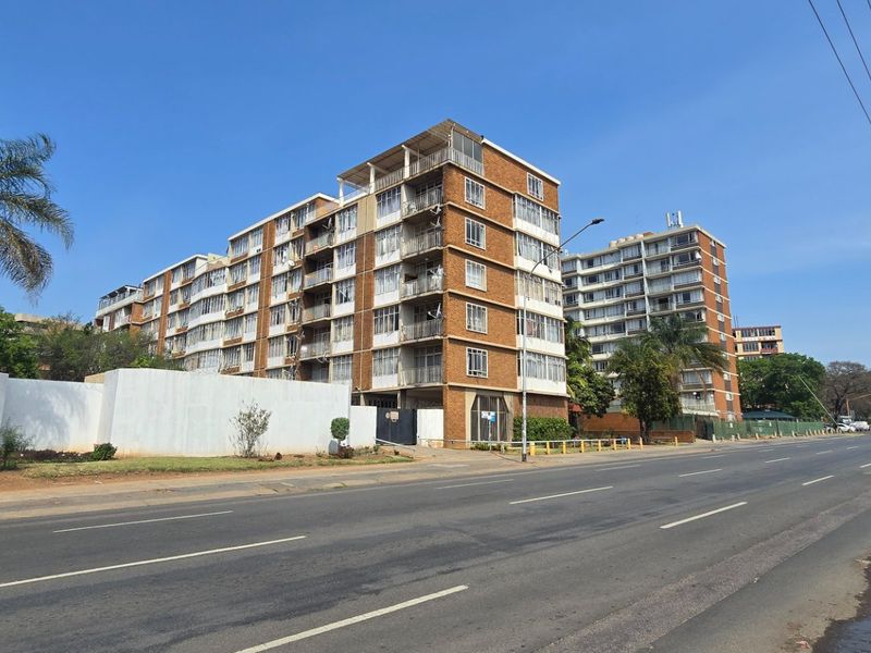 A street-level view of a multi-story apartment complex featuring brick facades and private balconies, situated along a main road.