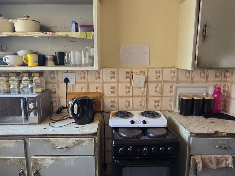 A dated kitchen featuring original cabinetry, tiled backsplash, and a freestanding stove, requiring significant cosmetic updates and modernization of fixed finishes.