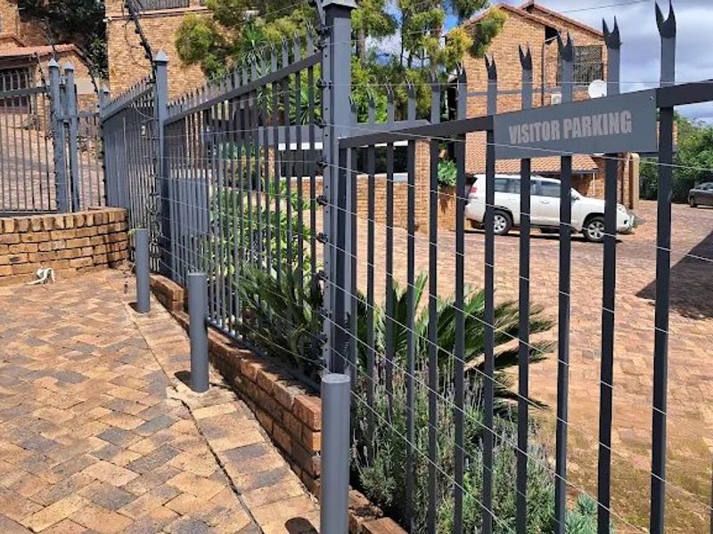 Front exterior view of the townhouse complex featuring secure steel fencing with electric wires, brick retaining walls, and designated visitor parking area.