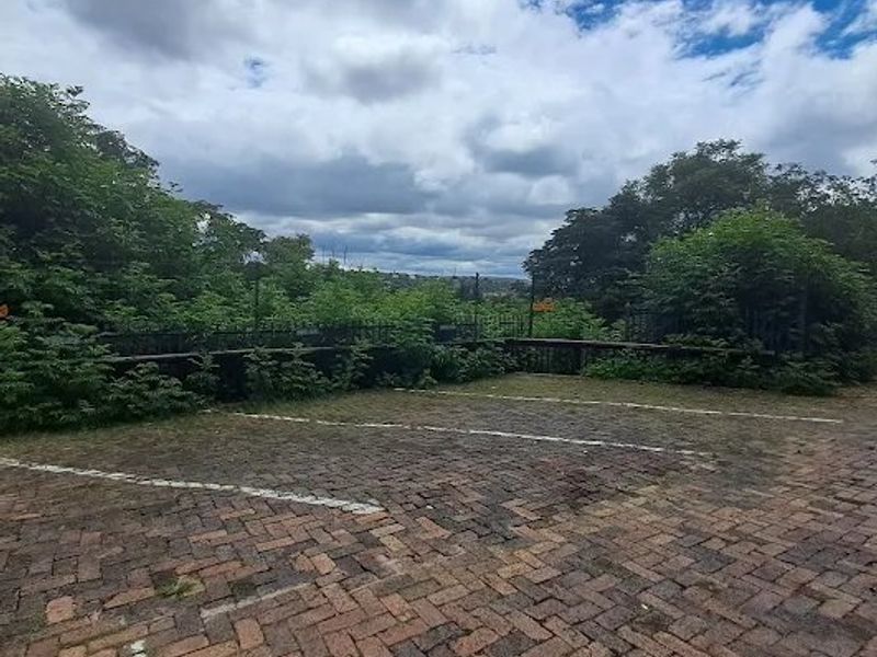 An outdoor paved parking area featuring marked bays, perimeter fencing, and a retaining wall, surrounded by established greenery.