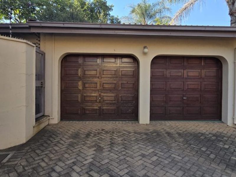 A double garage with wooden panel doors and a paved driveway, featuring exterior security fencing and wall-mounted lighting.