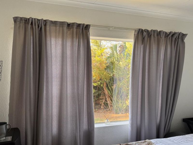 A bedroom featuring a window with a curtain rod, offering a view of the garden greenery outside.