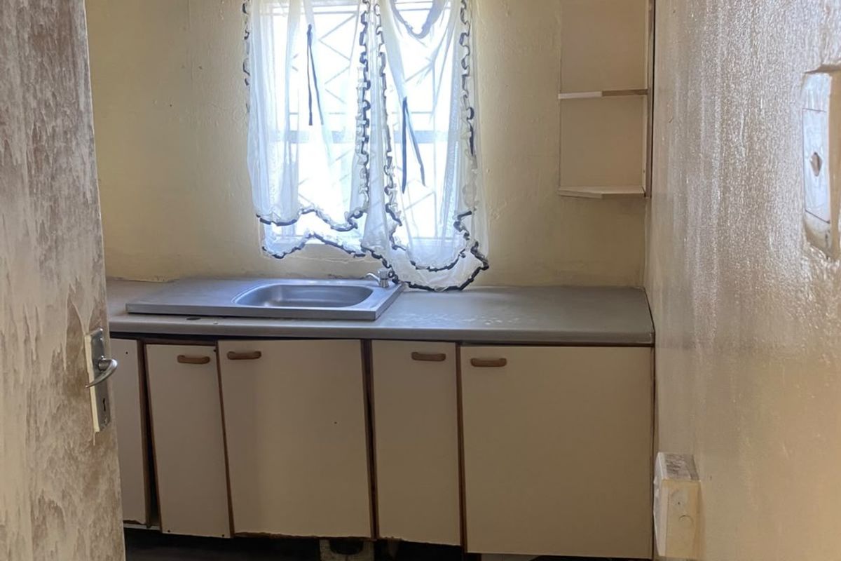 A small, dated kitchen area featuring basic cabinetry, a stainless steel sink, and built-in shelving, requiring significant cosmetic updates and renovation.
