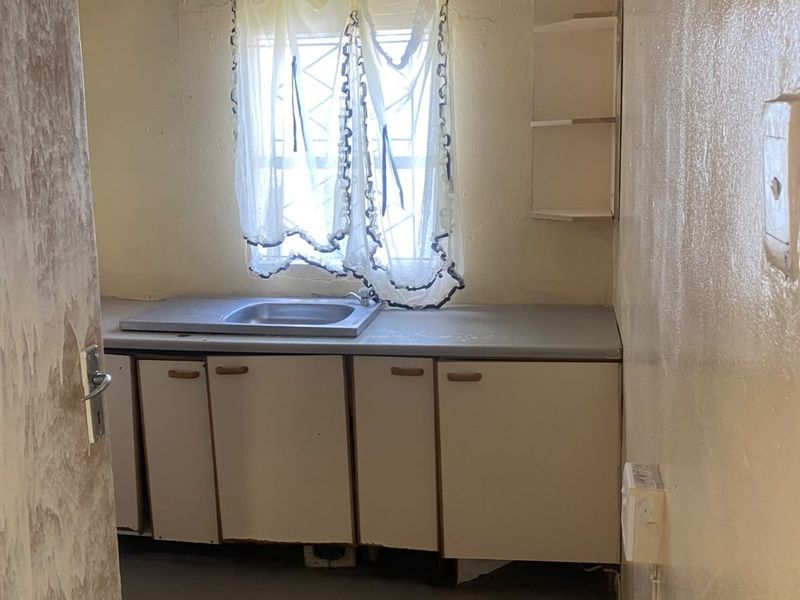 A small, dated kitchen area featuring basic cabinetry, a stainless steel sink, and built-in shelving, requiring significant cosmetic updates and renovation.