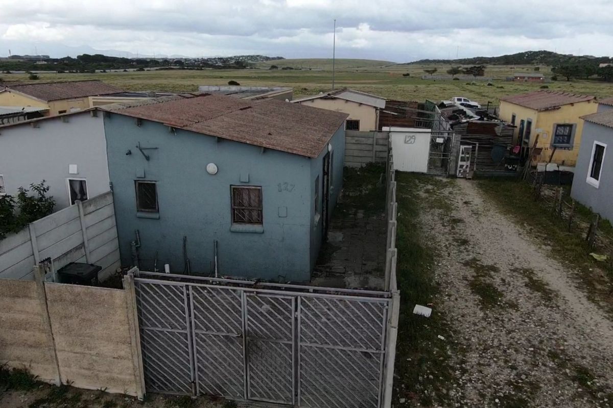 Aerial view of a residential property featuring a blue exterior, corrugated metal roof, and a fenced yard with an unpaved driveway.