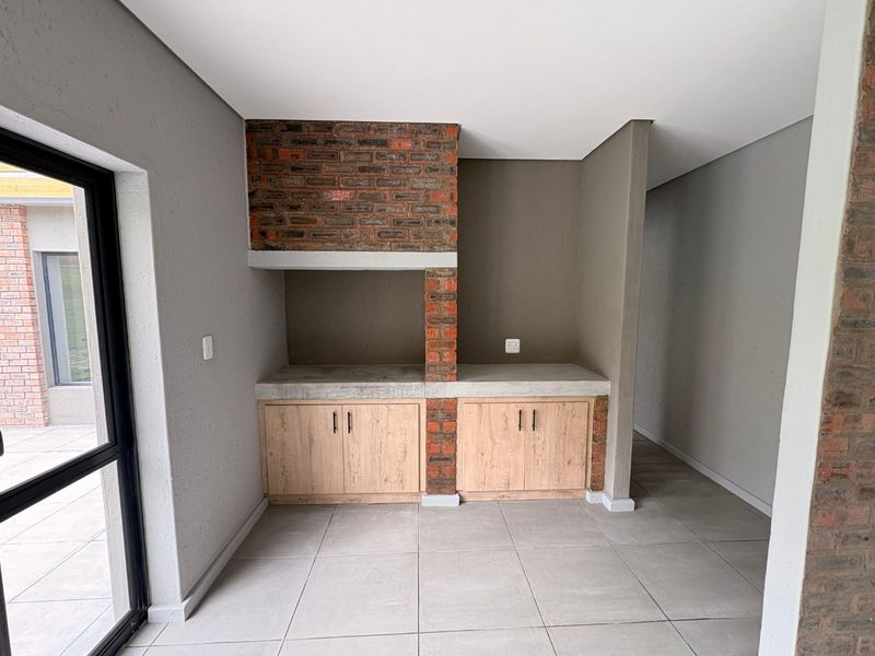 A modern covered patio area featuring a built-in brick braai, concrete prep surfaces, and light wood-finish cabinetry, opening onto an outdoor space.