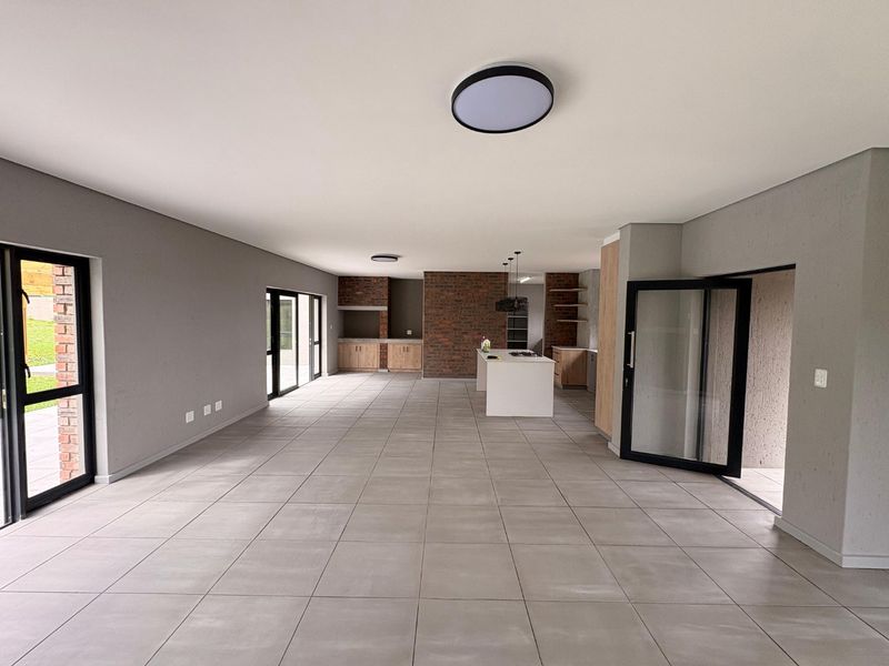 Spacious open-plan living and kitchen area featuring large format floor tiles, an exposed brick feature wall, built-in braai, and modern kitchen cabinetry.