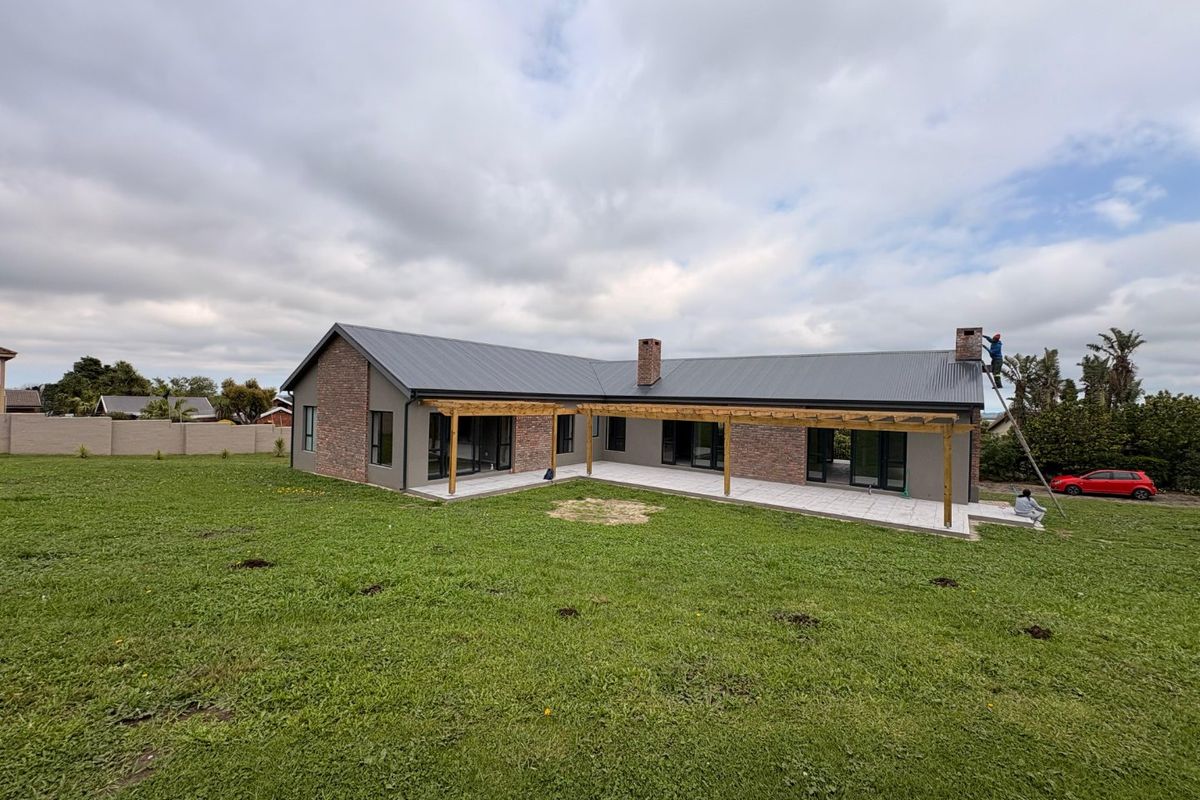 Rear view of a modern single-story home featuring a metal roof, brick accents, a timber pergola, and a paved patio overlooking a large lawn.