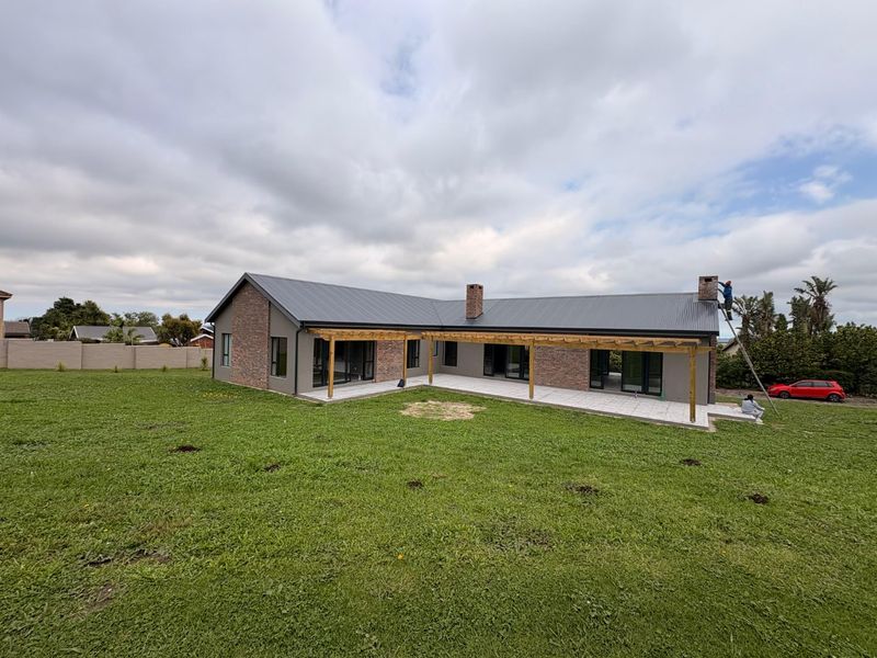 Rear view of a modern single-story home featuring a metal roof, brick accents, a timber pergola, and a paved patio overlooking a large lawn.