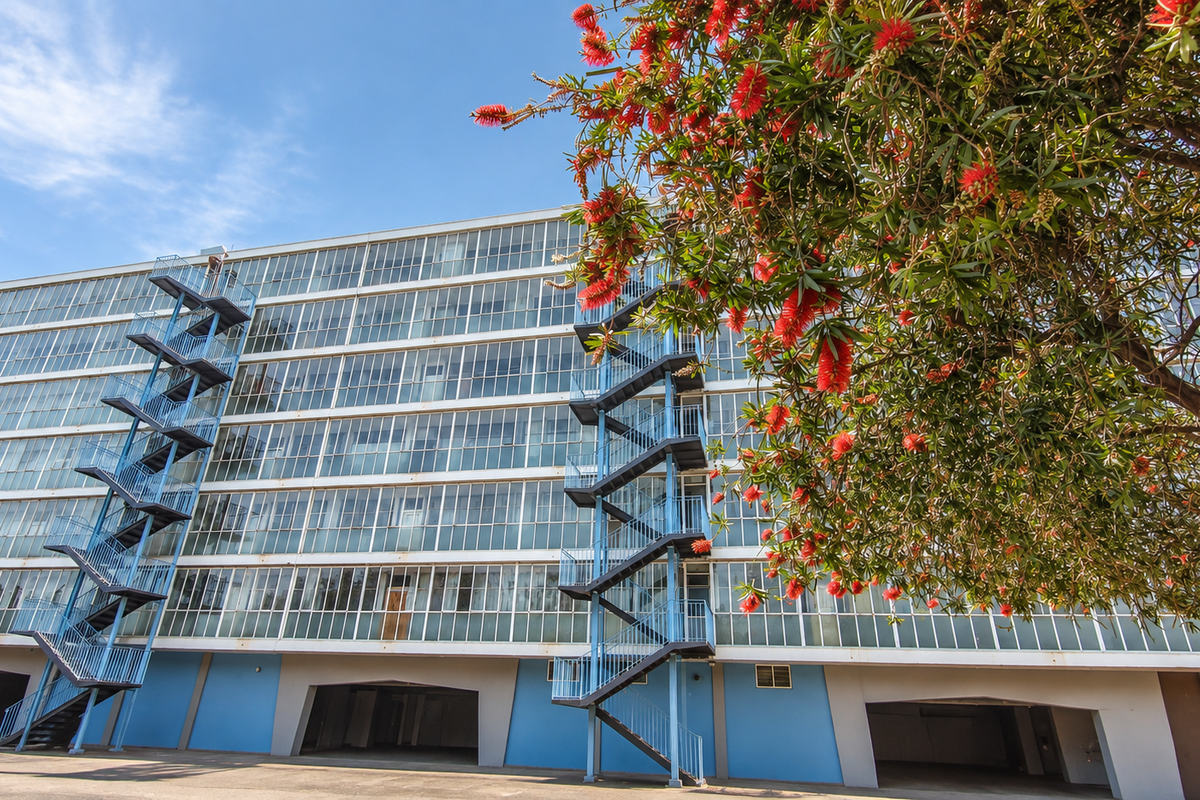 Exterior view of a multi-story apartment building featuring extensive glass window facades, external steel fire escape staircases, and ground-level covered parking.