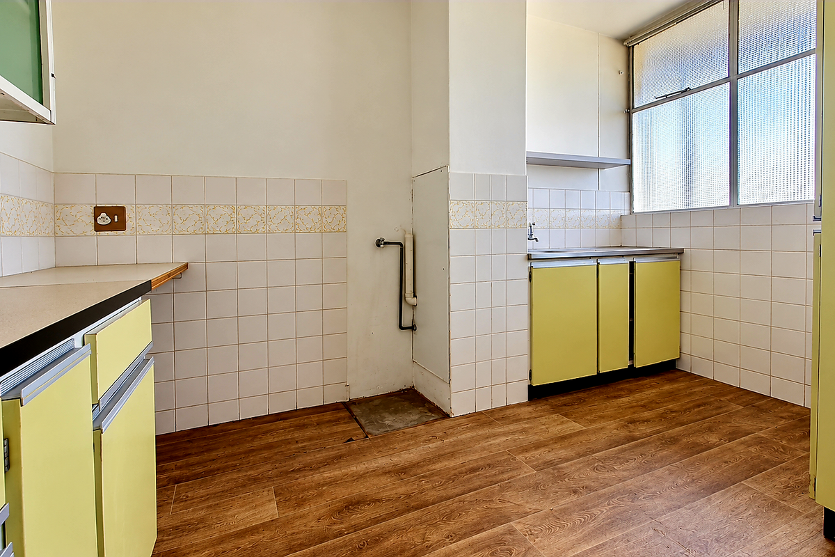 An empty, dated kitchen featuring original yellow cabinetry, tiled splashbacks, laminate countertops, and wood-look flooring, offering a blank canvas for modern renovation.