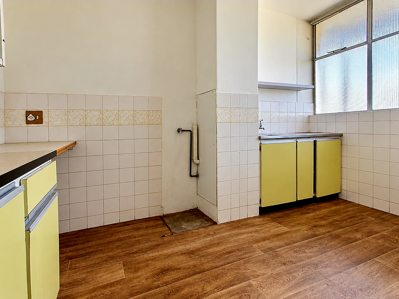 An empty, dated kitchen featuring original yellow cabinetry, tiled splashbacks, laminate countertops, and wood-look flooring, offering a blank canvas for modern renovation.