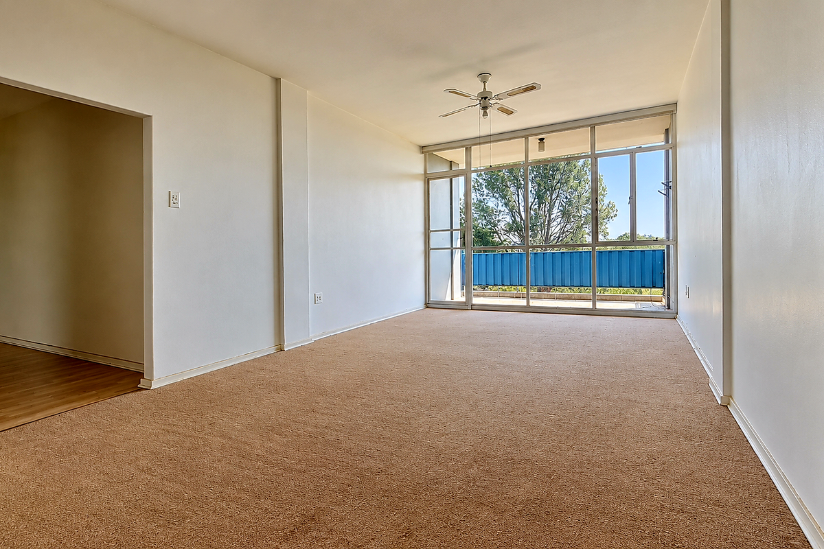 Spacious, empty living area featuring neutral carpeting, a ceiling fan, and large glass sliding doors providing natural light and access to the balcony.