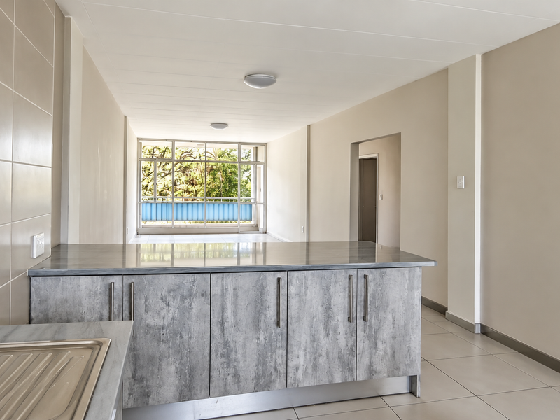 A bright, open-plan living and kitchen area featuring modern grey cabinetry, stone countertops, tiled flooring, and large windows allowing for ample natural light.