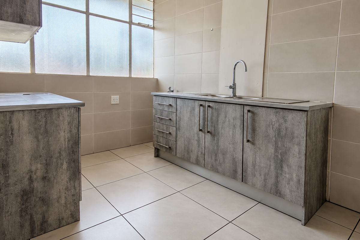A clean, modern kitchen featuring grey textured cabinetry, stainless steel sink, and neutral wall and floor tiling with ample natural light.