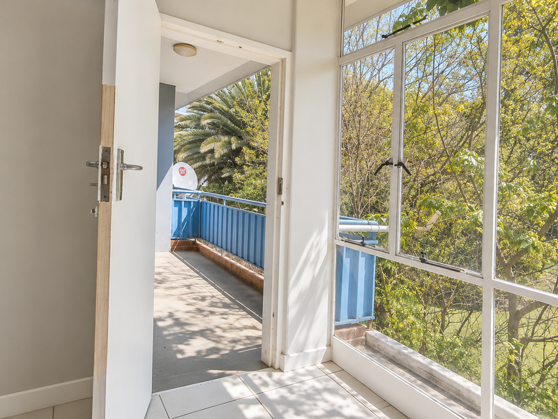 A bright, empty balcony area featuring tiled flooring, a metal balustrade, and large windows overlooking lush greenery.