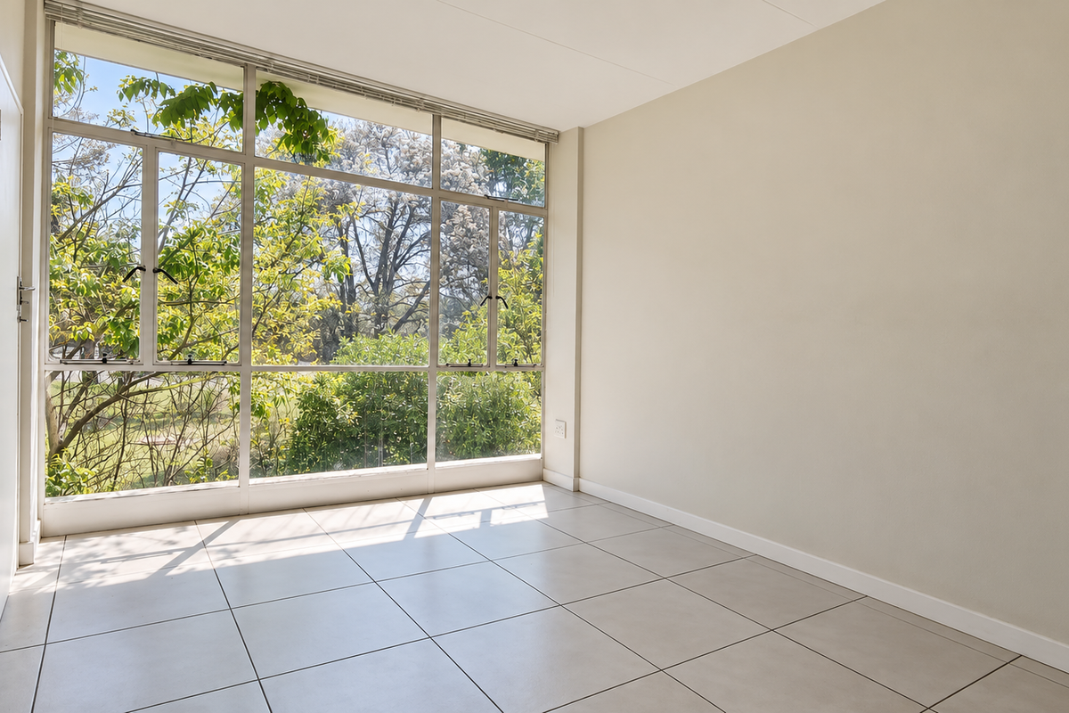 A bright, empty bedroom featuring large aluminium-framed windows that provide ample natural light and views of the surrounding greenery, finished with neutral tiled flooring.
