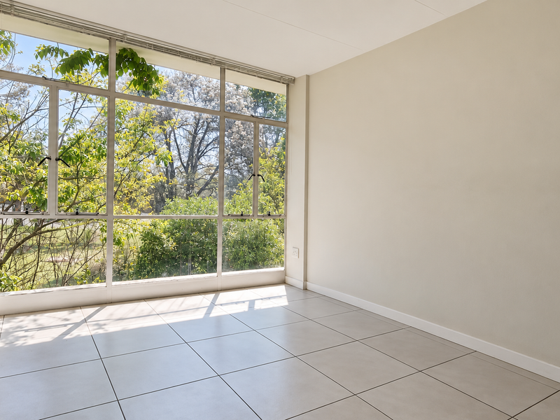 A bright, empty bedroom featuring large aluminium-framed windows that provide ample natural light and views of the surrounding greenery, finished with neutral tiled flooring.