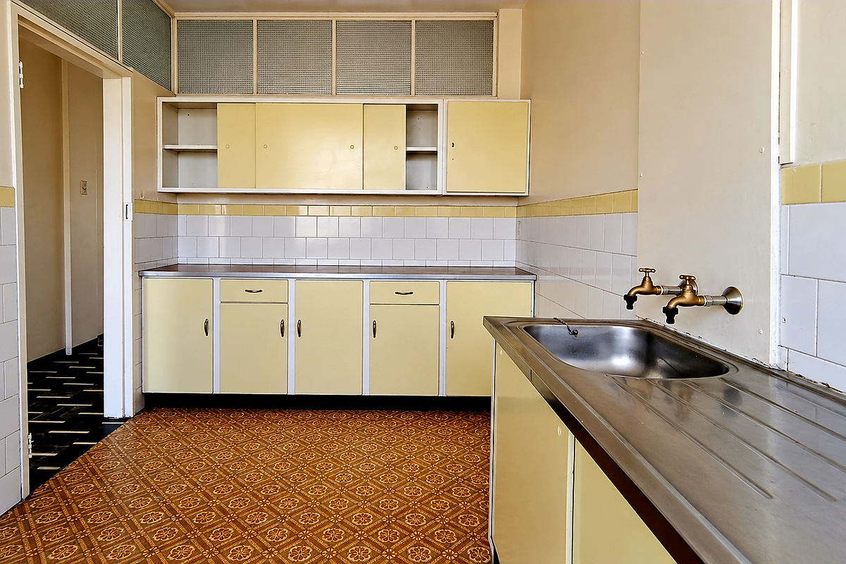 A dated, empty kitchen featuring original cabinetry, a stainless steel sink with wall-mounted taps, and classic patterned vinyl flooring.