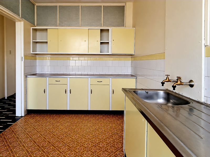 A dated, empty kitchen featuring original cabinetry, a stainless steel sink with wall-mounted taps, and classic patterned vinyl flooring.