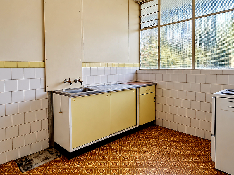 A dated, empty kitchen featuring original cabinetry, a stainless steel sink, tiled splashbacks, and a freestanding electric stove with large louvered windows.