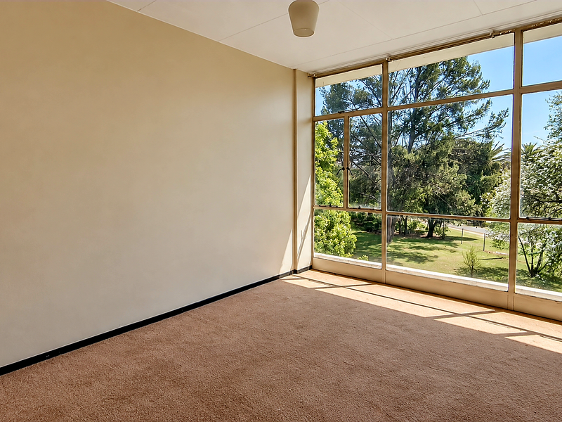 An empty, sunlit living area featuring expansive floor-to-ceiling windows with pleasant garden views and neutral wall finishes.