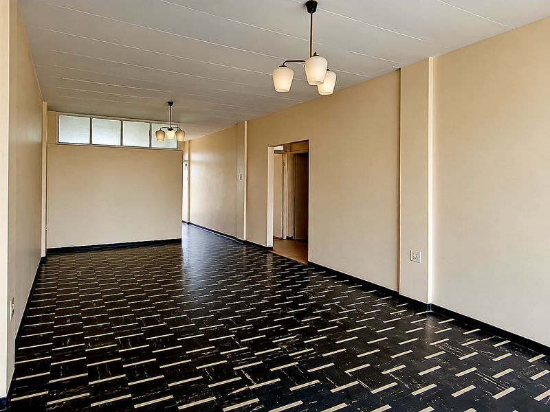 A spacious, empty living area featuring neutral walls, dark patterned vinyl flooring, and classic pendant lighting fixtures.
