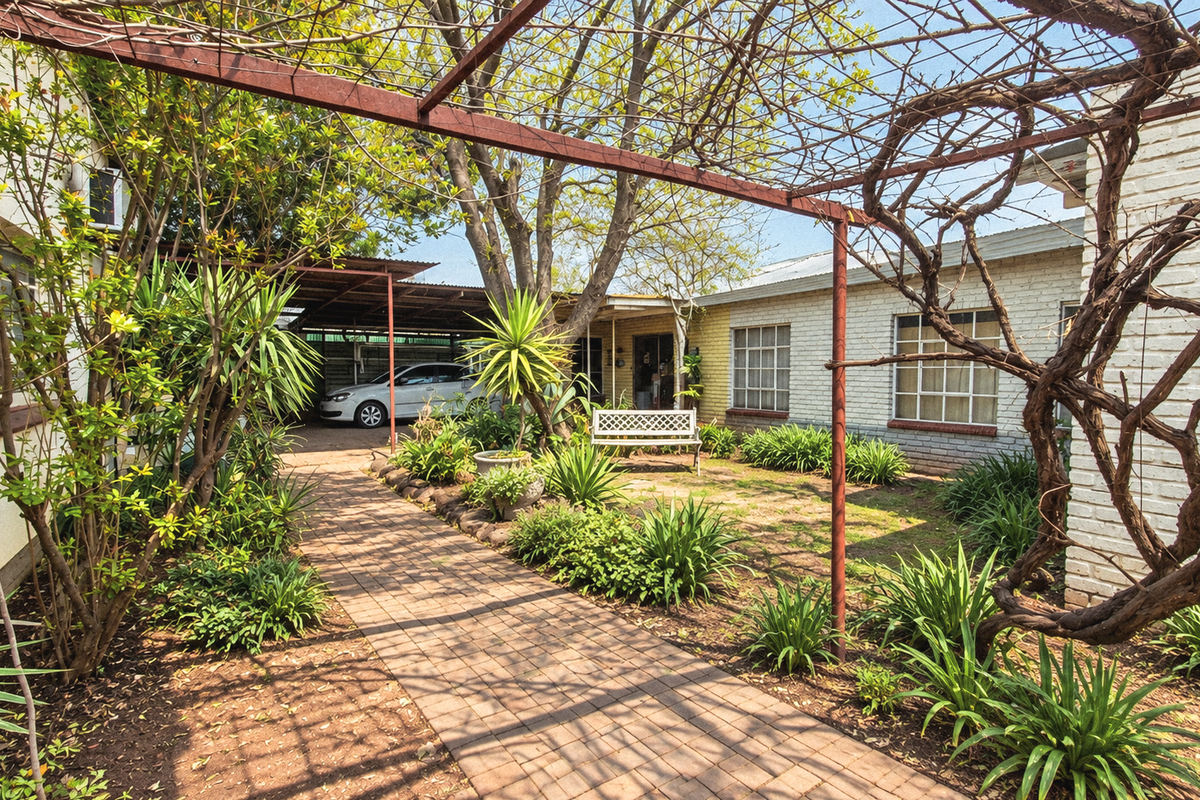 A rear garden area featuring a brick paved pathway, a metal pergola structure, and established landscaping alongside the painted brick exterior of the house.
