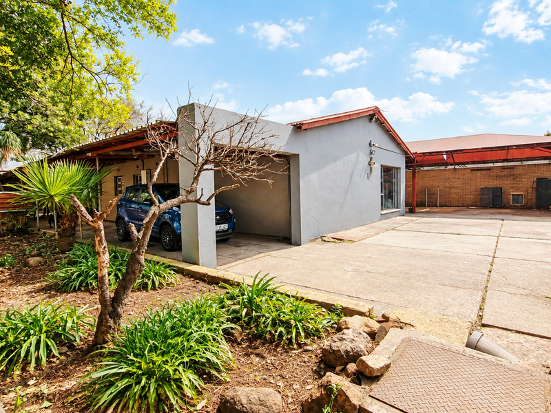 Rear exterior view featuring a large concrete driveway, covered carport area, and brick boundary walling, providing ample parking space.