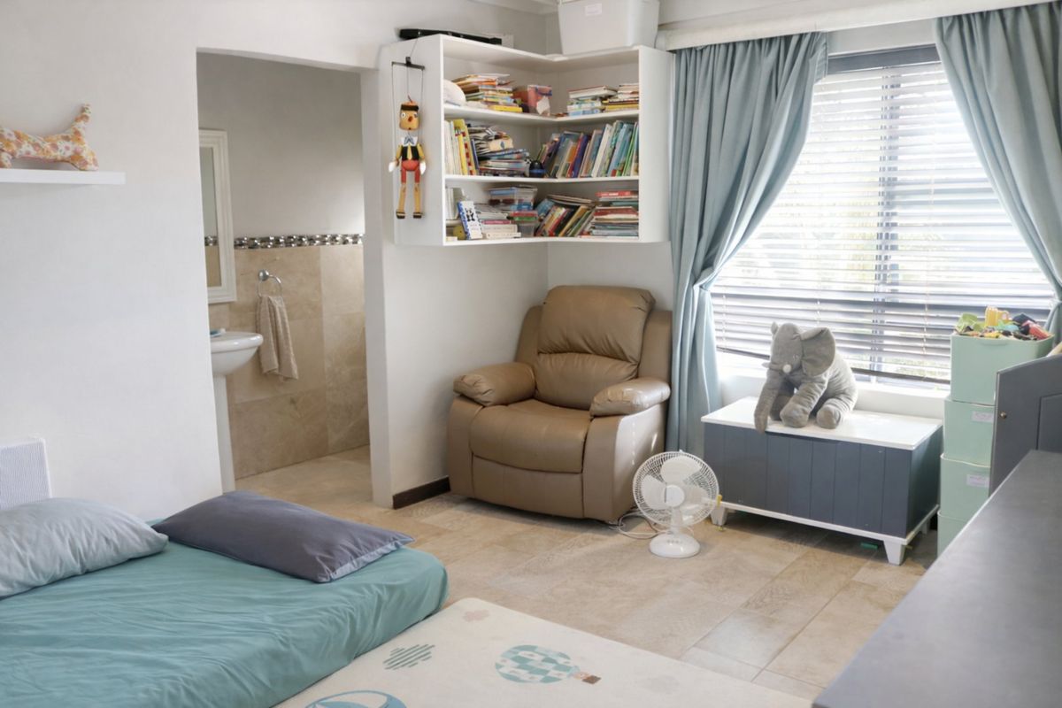 A bedroom featuring tiled flooring, built-in corner shelving, and an en-suite bathroom, with large windows fitted with venetian blinds.