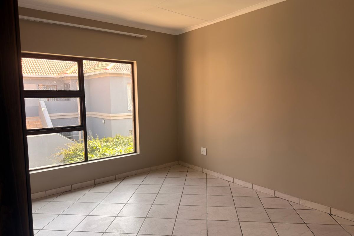 A bright, empty bedroom featuring neutral wall tones, tiled flooring, and a large aluminium-framed window providing natural light.