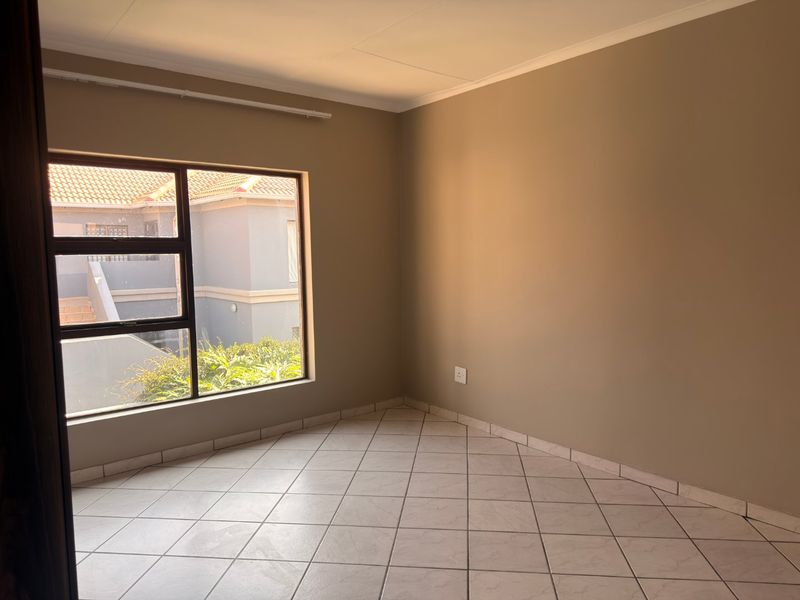 A bright, empty bedroom featuring neutral wall tones, tiled flooring, and a large aluminium-framed window providing natural light.