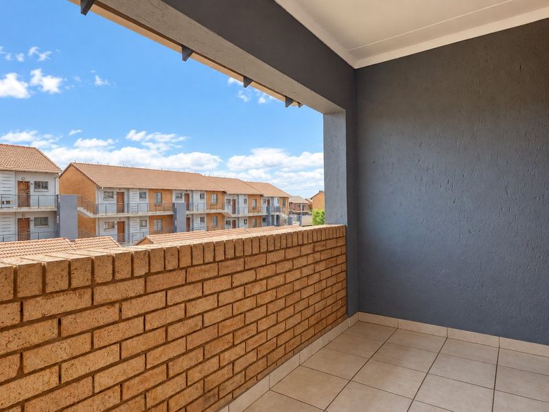 A clean, covered balcony featuring a brick balustrade, tiled flooring, and a neutral grey painted wall, offering an open view of the complex.