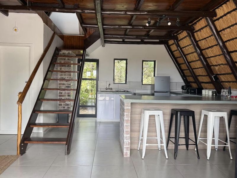 Open plan living area featuring a kitchen island, exposed timber beams, thatched roof detail, and a timber staircase leading to an upper level.