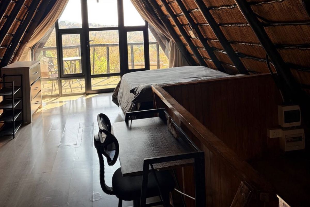 A cozy loft-style bedroom featuring a traditional thatched roof with exposed timber rafters, laminate flooring, and large glass doors leading to a balcony.
