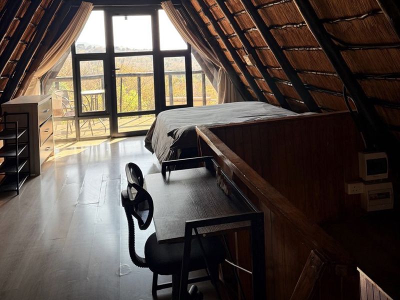 A cozy loft-style bedroom featuring a traditional thatched roof with exposed timber rafters, laminate flooring, and large glass doors leading to a balcony.
