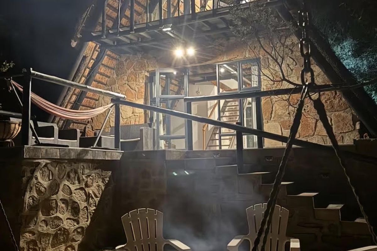 A charming A-frame house at night, featuring stone exterior walls, a thatched roof, a balcony, and a stone-paved patio area with a fire pit.