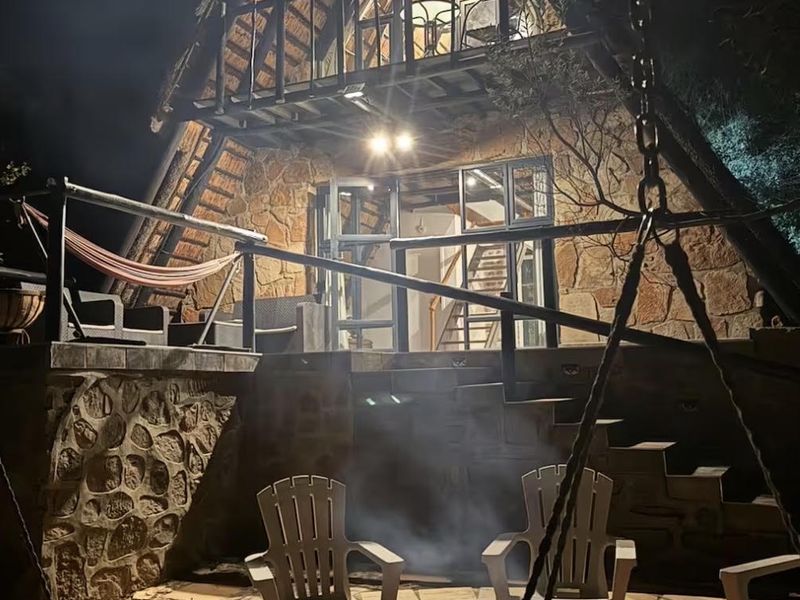 A charming A-frame house at night, featuring stone exterior walls, a thatched roof, a balcony, and a stone-paved patio area with a fire pit.