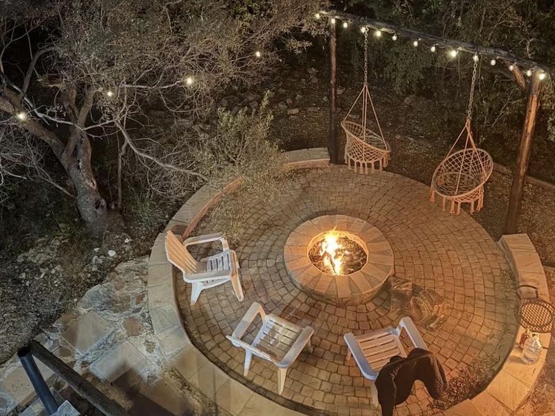 An inviting outdoor garden area featuring a circular stone-paved fire pit, overhead timber beam structure with string lighting, and natural landscaping.