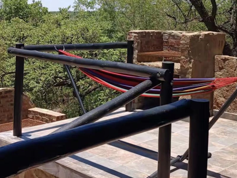 An elevated outdoor patio featuring stone masonry, tiled flooring, and a built-in stone braai, offering a scenic view of the surrounding trees.